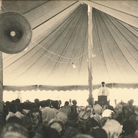 men and women listening, under a large tent. Listening to a speaker